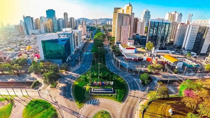 Fotografia aérea de Barueri mostrando uma grande avenida que se divide em torno de um canteiro central arborizado, cercada por prédios comerciais altos e modernos sob um céu claro.
